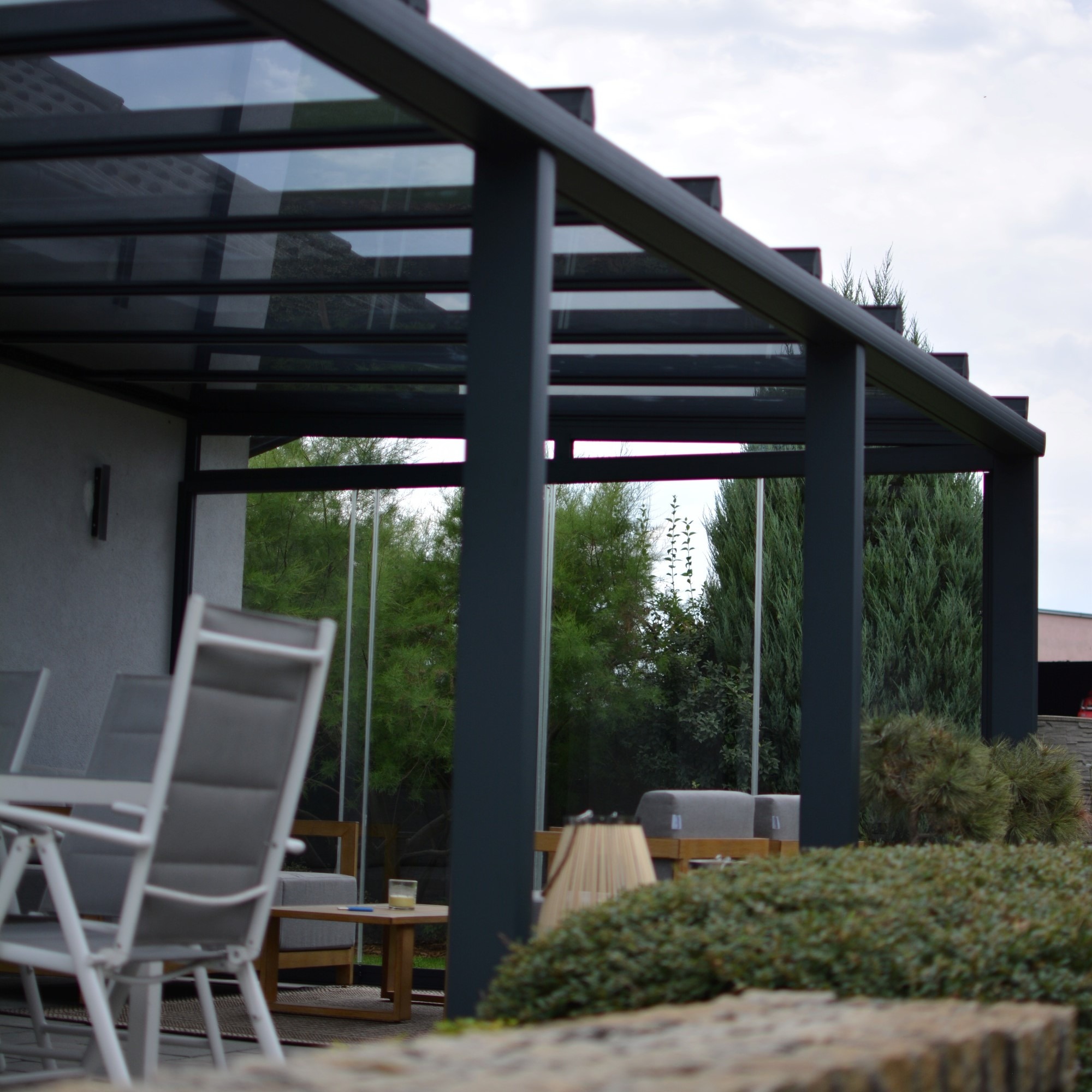 Nowoczesna pergola z ciemnego metalu z przeszklonym dachem, widok z zewnątrz na meble ogrodowe i zieleń. Minimalistyczny design.