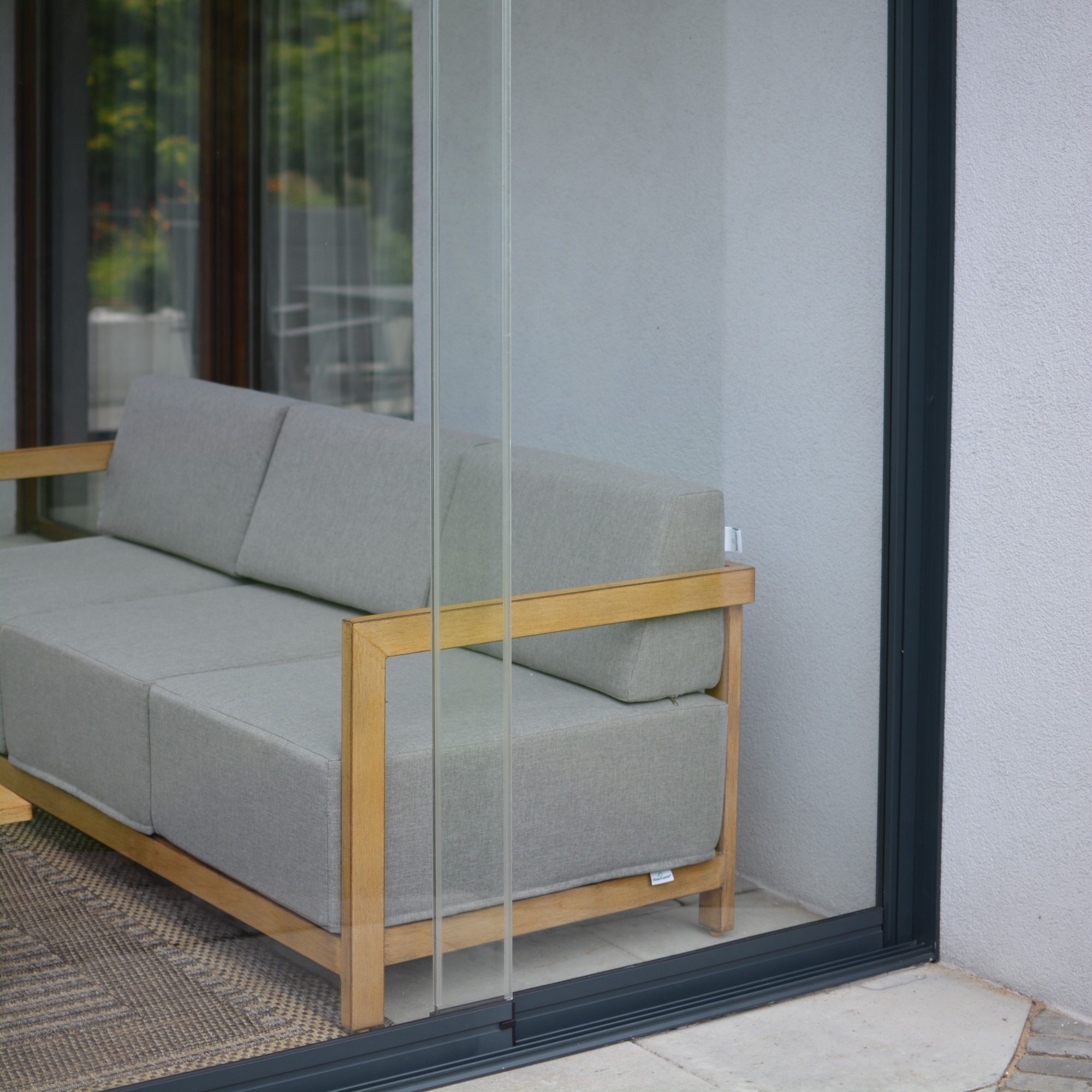Nowoczesna zabudowa balkonu z przesuwnymi szklanymi panelami i drewnianym narożnikiem z szarą tapicerką. Widok z zewnątrz, minimalistyczny design.