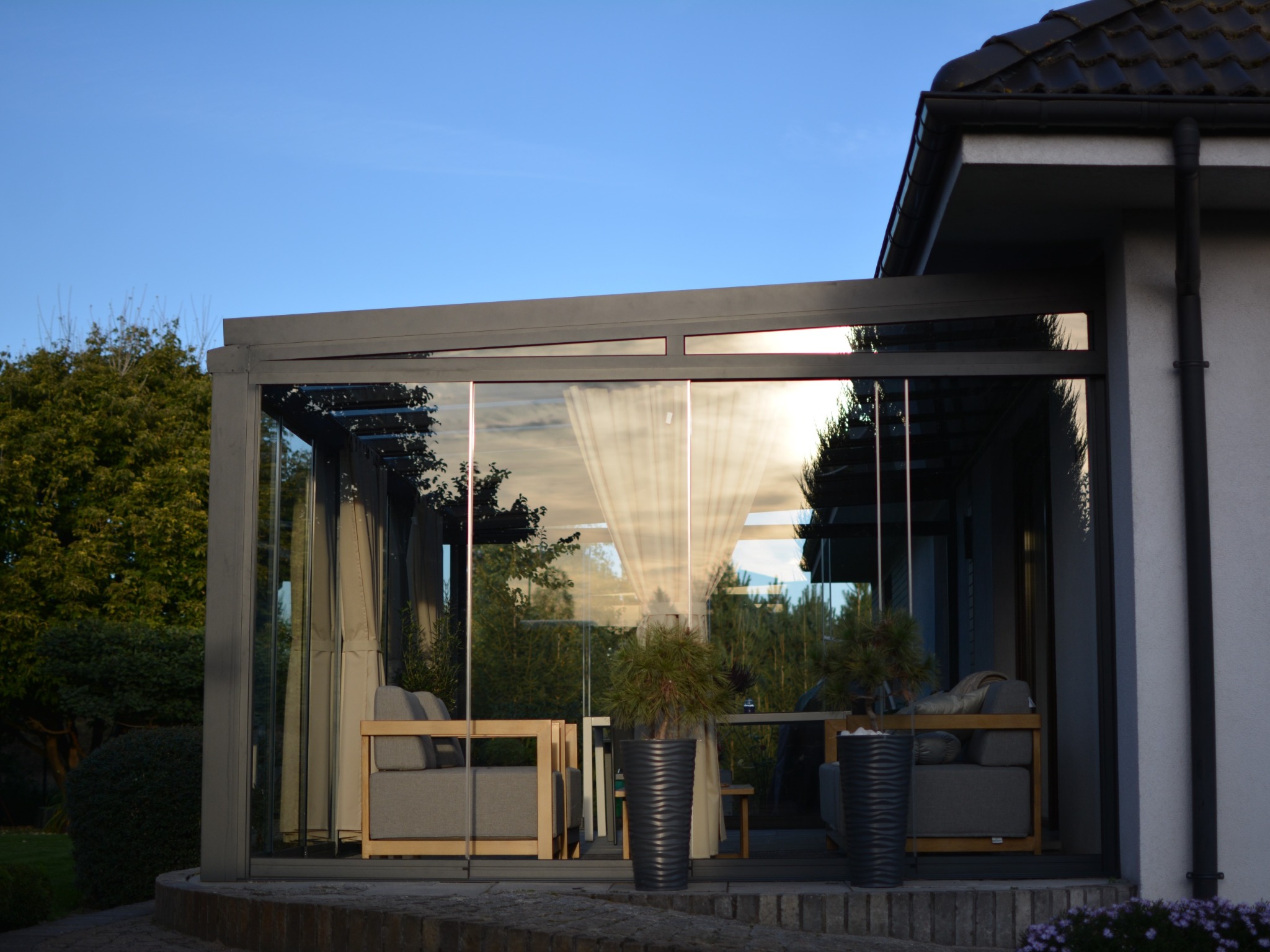 Szklana zabudowa tarasu z szarą ramą, widoczne meble wypoczynkowe i rośliny w donicach. W tle zieleń ogrodu i błękitne niebo. Nowoczesny design.