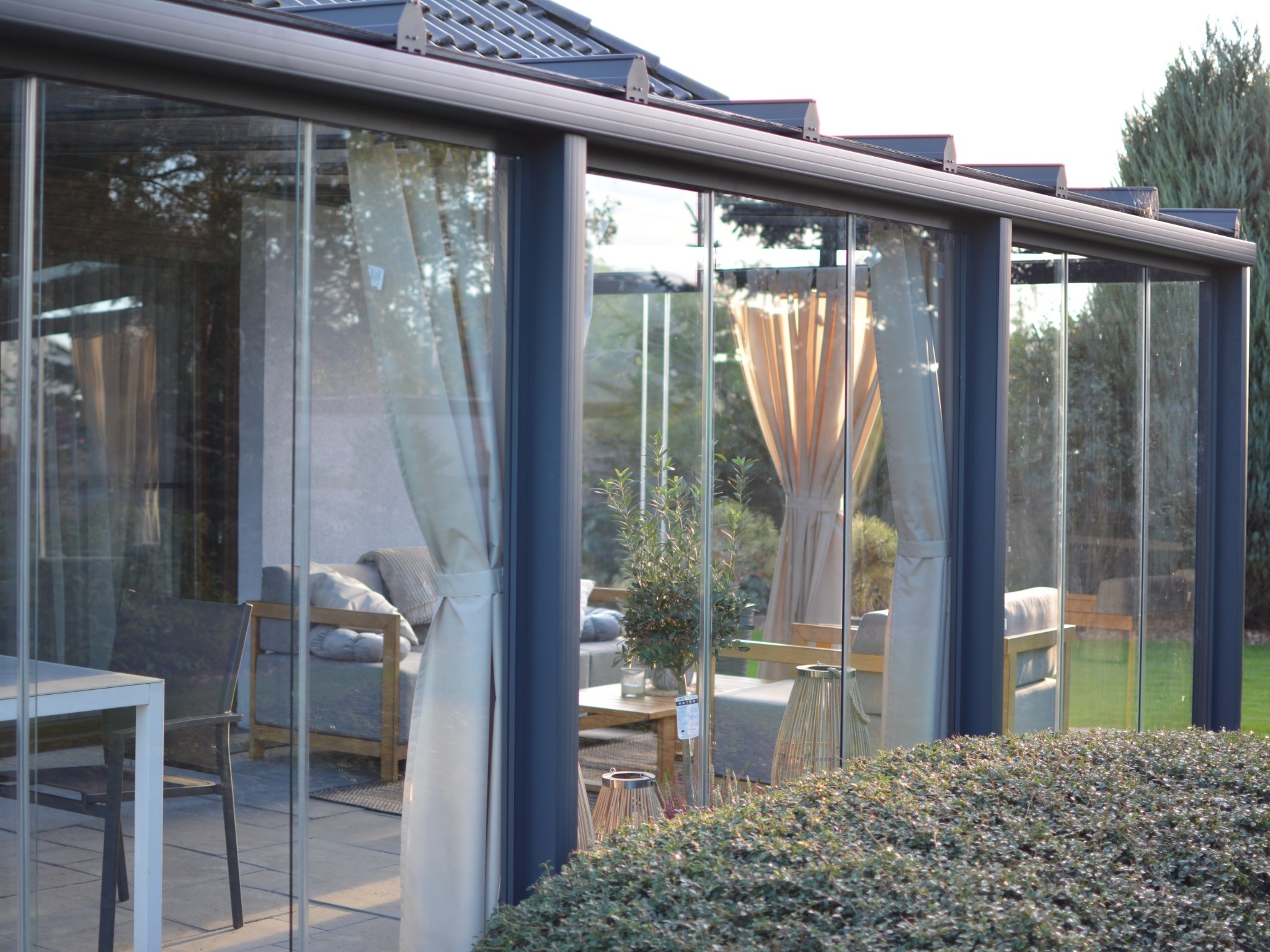 Szklana zabudowa tarasu z ciemną ramą, widok z zewnątrz na meble ogrodowe i dekoracje. Częściowo zasłonięte firankami, zieleń w tle.