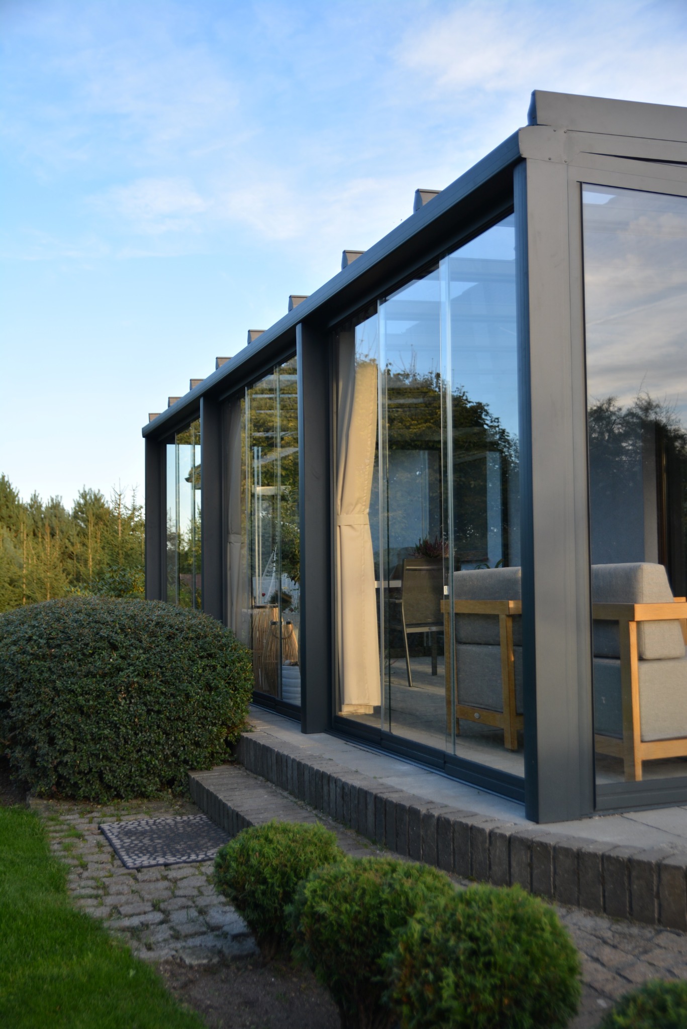 Szklana zabudowa tarasu z szarymi ramami, widok z zewnątrz. Wnętrze z meblami ogrodowymi, zasłony. Otoczenie: kostka brukowa, krzewy, drzewa w tle. Ujęcie z dołu.