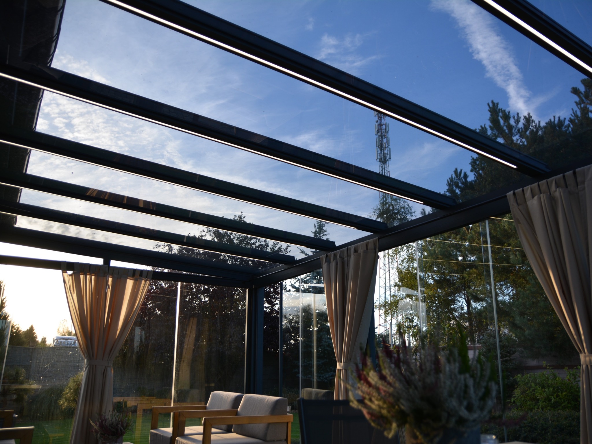 Nowoczesna zabudowa balkonu z metalową konstrukcją i szklanym dachem, oświetlona listwami LED. Widok z wnętrza w Czarnkowie.