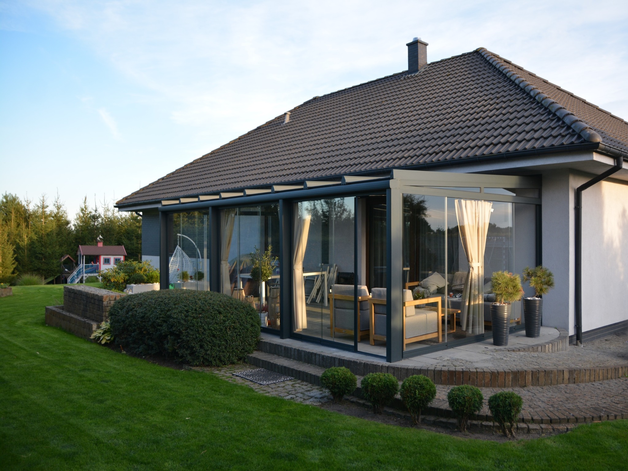 Szklana zabudowa balkonu z przesuwnymi panelami, widok z zewnątrz na taras z meblami ogrodowymi i zadbanym trawnikiem. Nowoczesny design, jasne wnętrze.