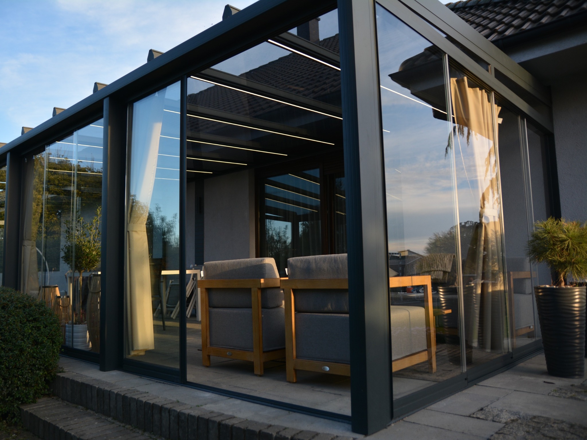 Szklana zabudowa tarasu z szarymi ramami, oświetleniem LED i meblami wypoczynkowymi wewnątrz. Widoczne rośliny w donicach i fragment domu w tle.