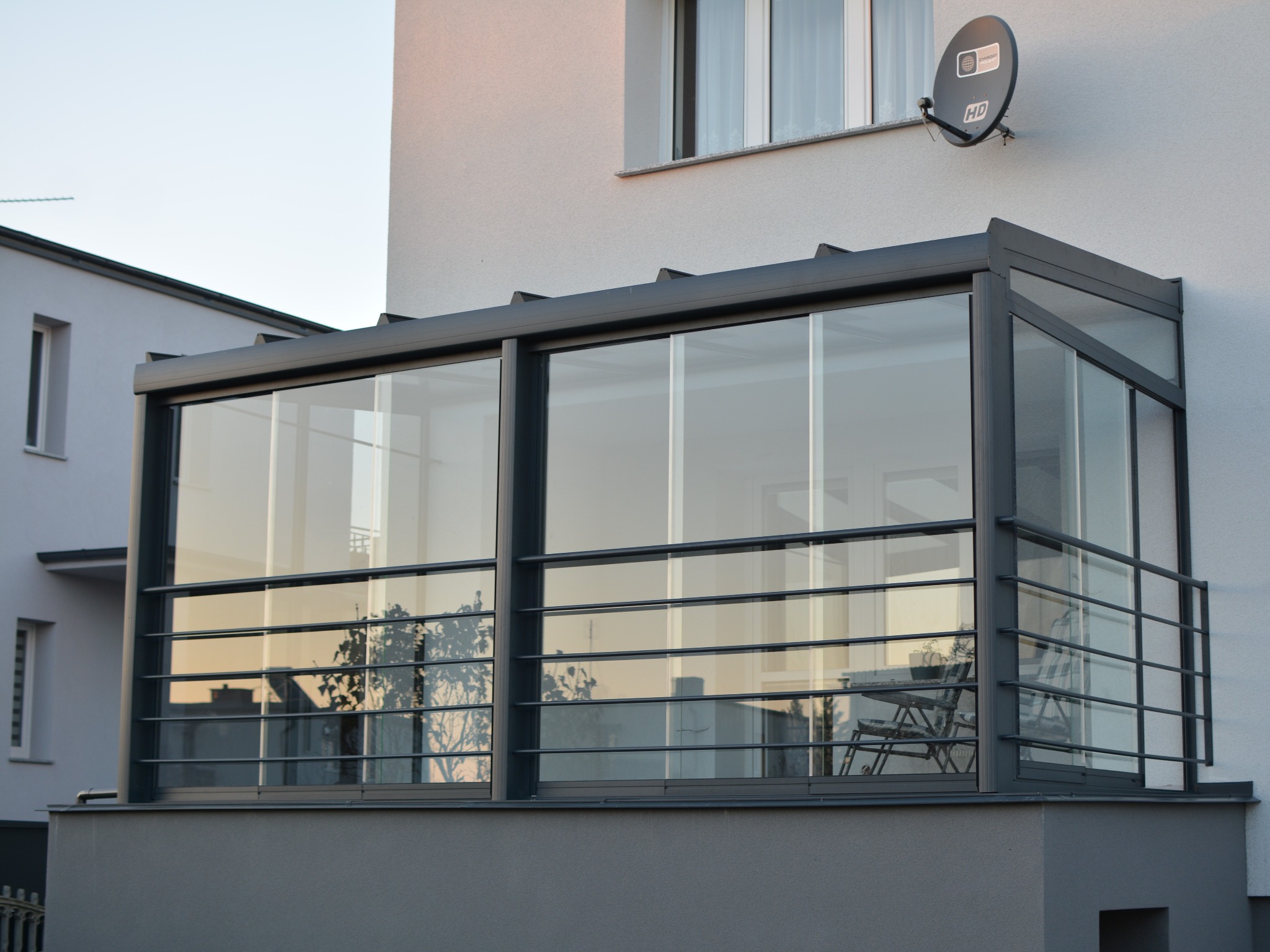 Szklana zabudowa balkonu z metalową ramą i balustradą, widok z zewnątrz. W tle budynki mieszkalne i błękitne niebo. Nowoczesny design, minimalistyczny styl.