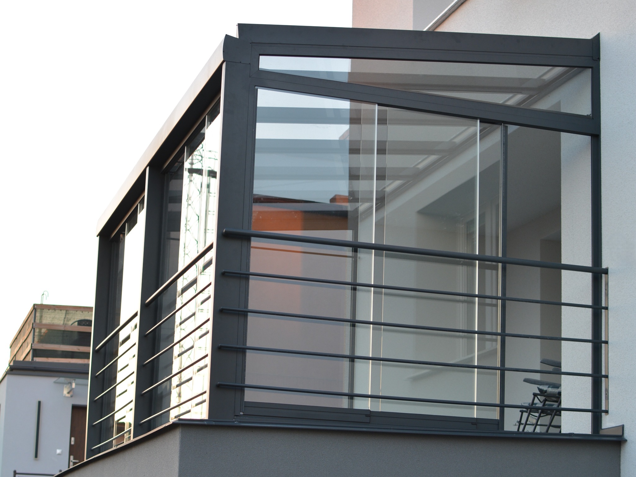 Nowoczesna zabudowa balkonu z przesuwnymi szklanymi panelami w grafitowej ramie, z widoczną metalową balustradą i elementami architektonicznymi budynku w tle.