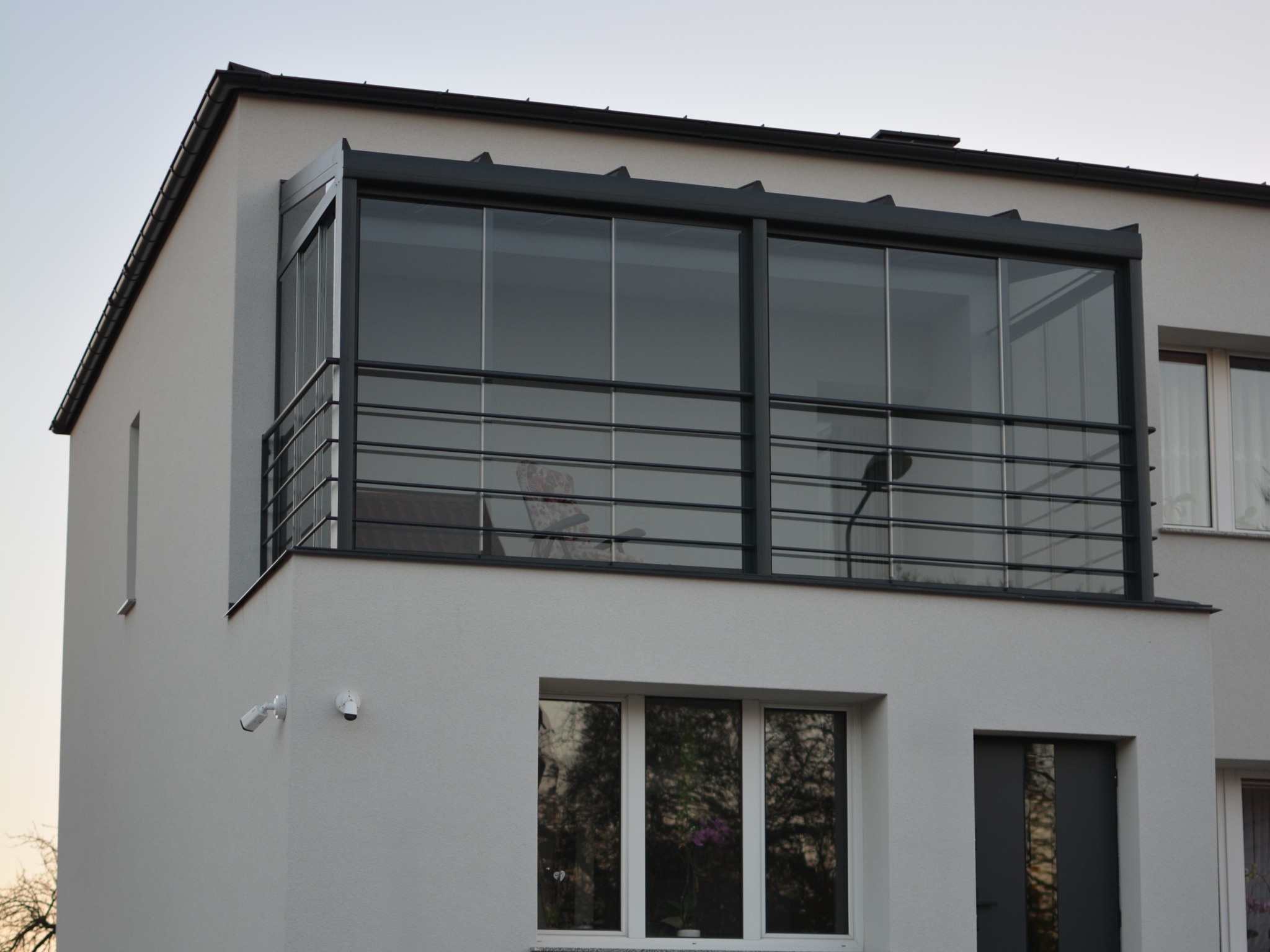 Nowoczesna zabudowa balkonu z czarną, metalową ramą i szklanymi panelami, widoczna z zewnątrz budynku. Minimalistyczny design, jasna elewacja, krzesło wewnątrz.