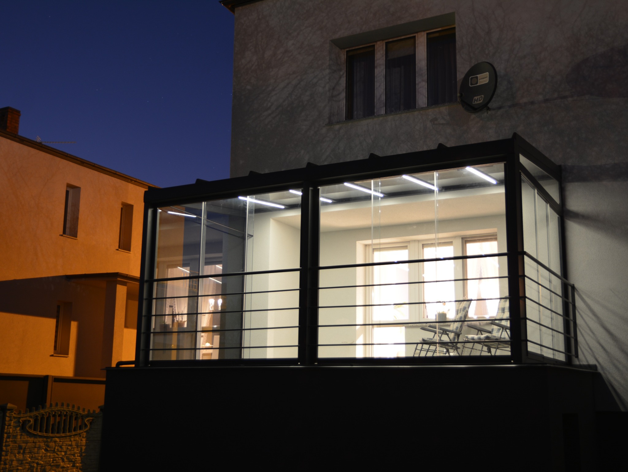 Szklana zabudowa balkonu z metalową ramą, oświetlona od wewnątrz, widoczna nocą. W tle budynki i ciemne niebo. Minimalistyczny design.