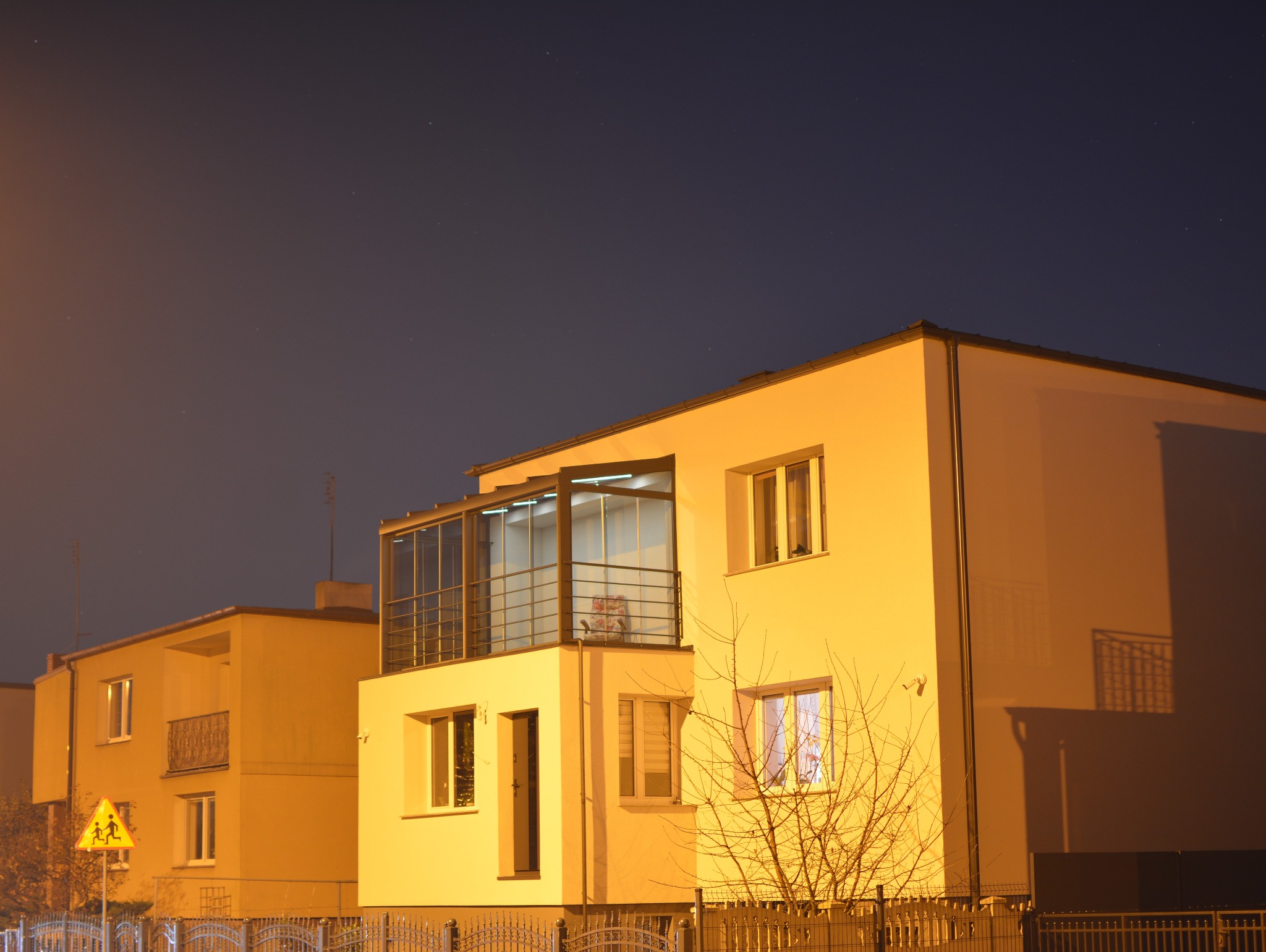 Nowoczesny dom z zabudowanym balkonem, oświetlony ciepłym światłem nocą. Widoczna metalowa balustrada i przeszklona konstrukcja zabudowy balkonu. W tle ciemne niebo.