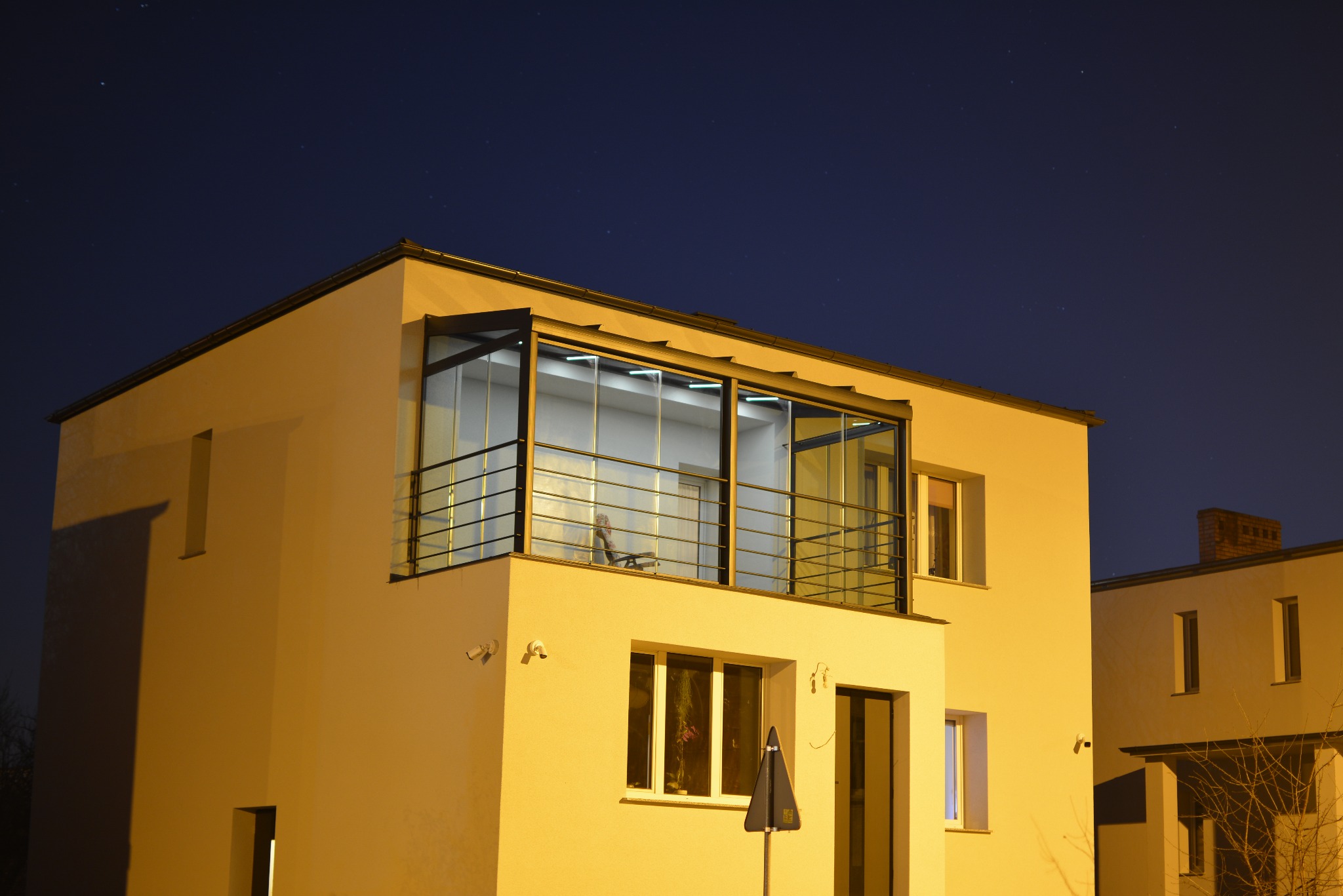 Nowoczesny budynek z zabudowanym balkonem, widok nocą. Szklana zabudowa z czarną ramą i metalową balustradą. Ciepłe oświetlenie elewacji kontrastuje z ciemnym niebem.