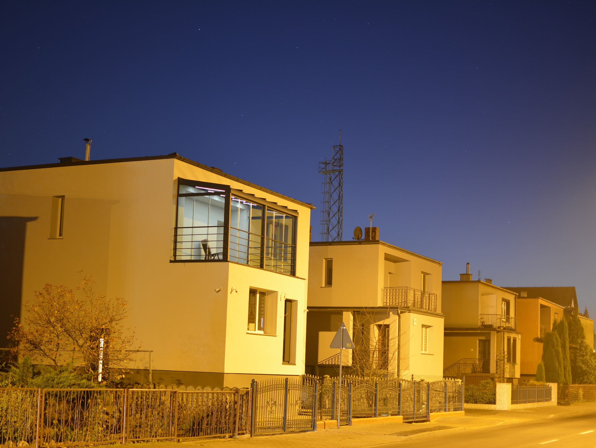 Nowoczesna zabudowa balkonu z metalową ramą i szklanymi panelami, widoczna nocą. W tle inne budynki i niebo z gwiazdami. Oświetlenie uliczne nadaje ciepły odcień.