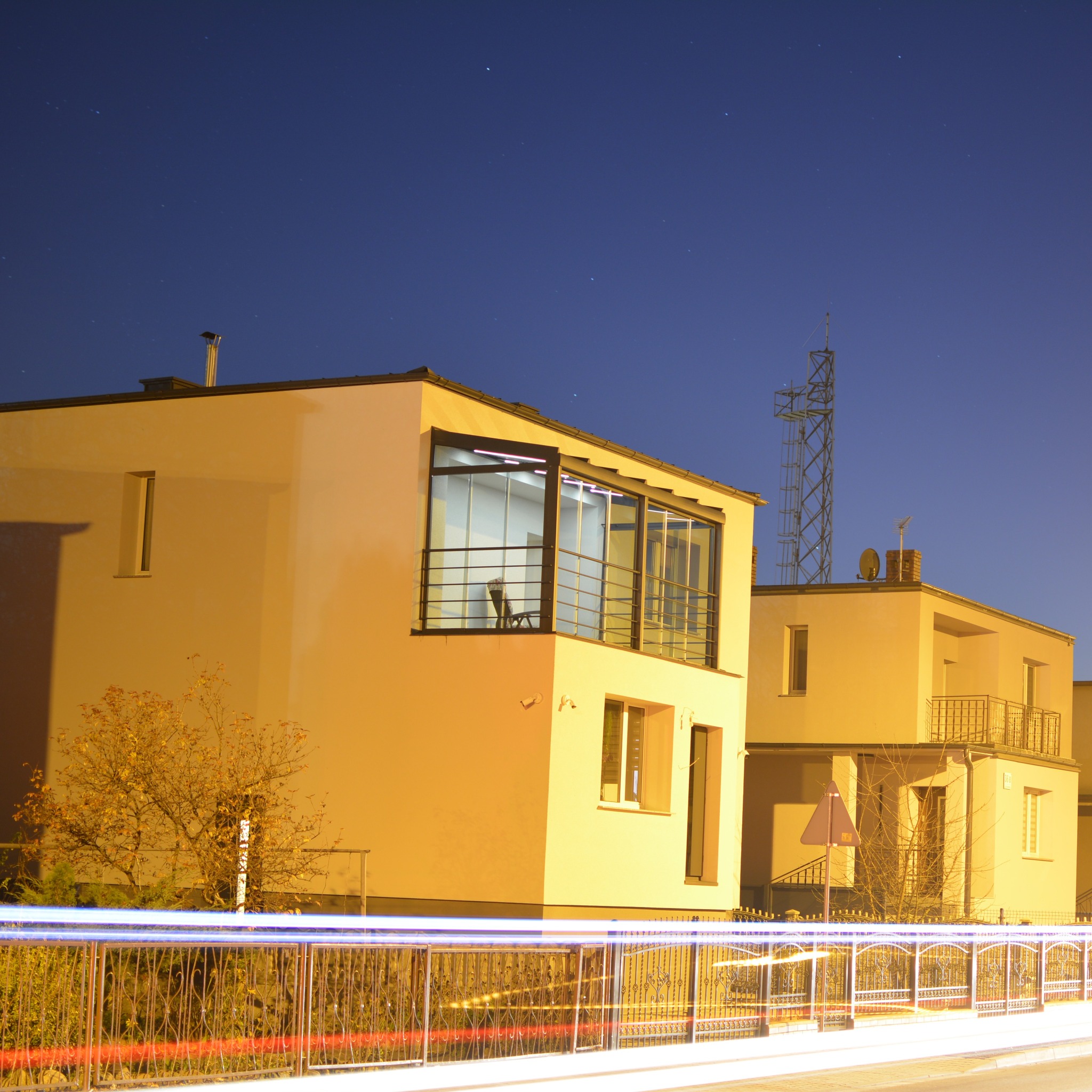 Nowoczesny dom z zabudowanym balkonem i metalowym ogrodzeniem, nocne ujęcie z smugami świateł przejeżdżających aut. Widoczna antena na dachu sąsiedniego budynku.