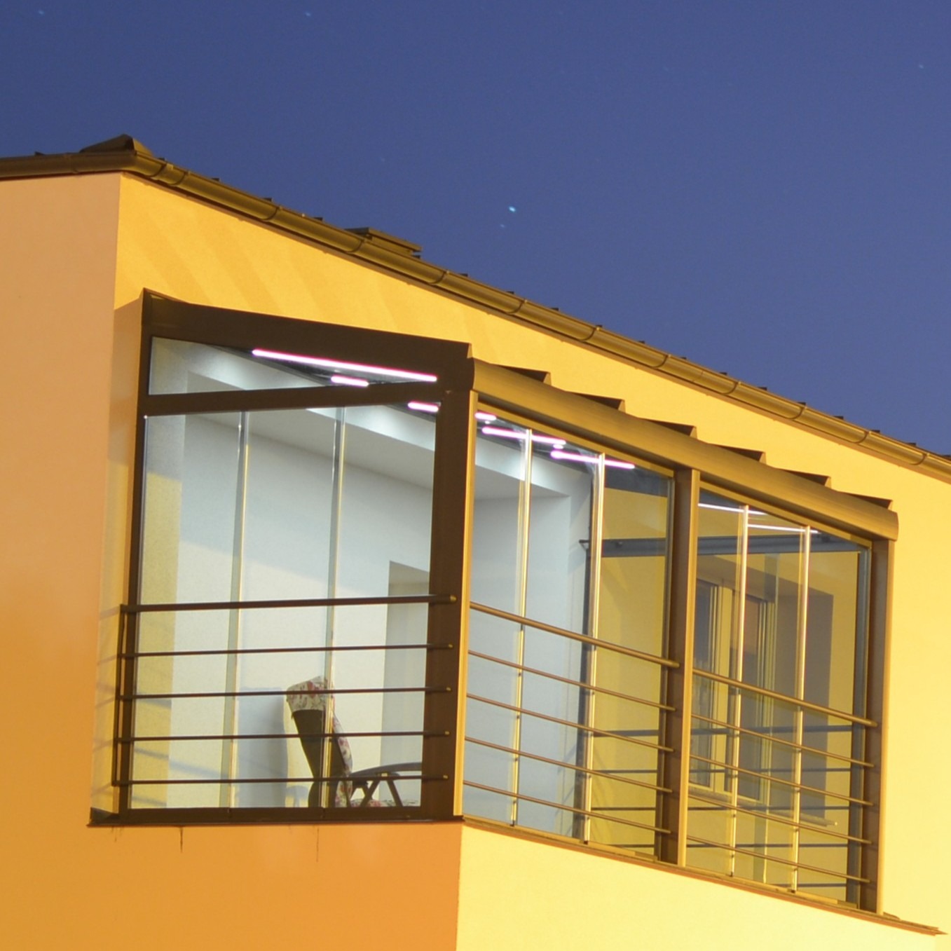 Nowoczesna zabudowa balkonu z metalową ramą i szklanymi panelami, oświetlona od wewnątrz, widoczna z zewnątrz budynku w nocnej scenerii. Minimalistyczny design.