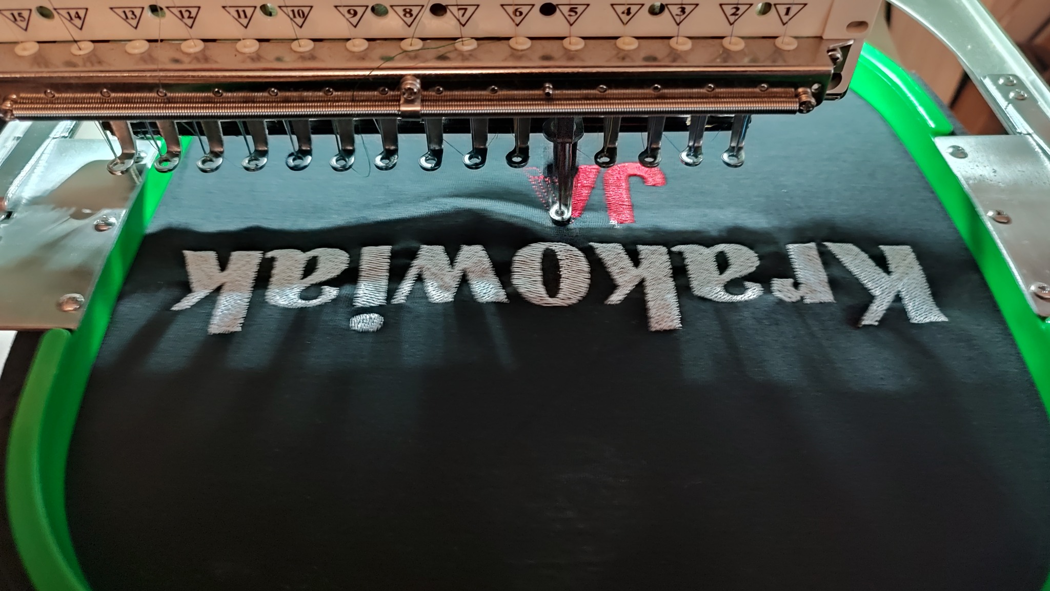 Proces tworzenia haftu na czarnej koszulce z napisem 'Krakowiak!' i inicjałami 'JT' w kolorze czerwonym, widoczna wieloigłowa maszyna do haftowania w trakcie pracy.