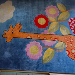 Dawdom Usługi Sprzątające - Niebieski dywan dziecięcy z motywem żyrafy, kwiatów i grzyba. Żyrafa ma pomarańczowe plamy z czerwonym obrysem. Dywan leży na podłodze, widoczny fragment ramy.