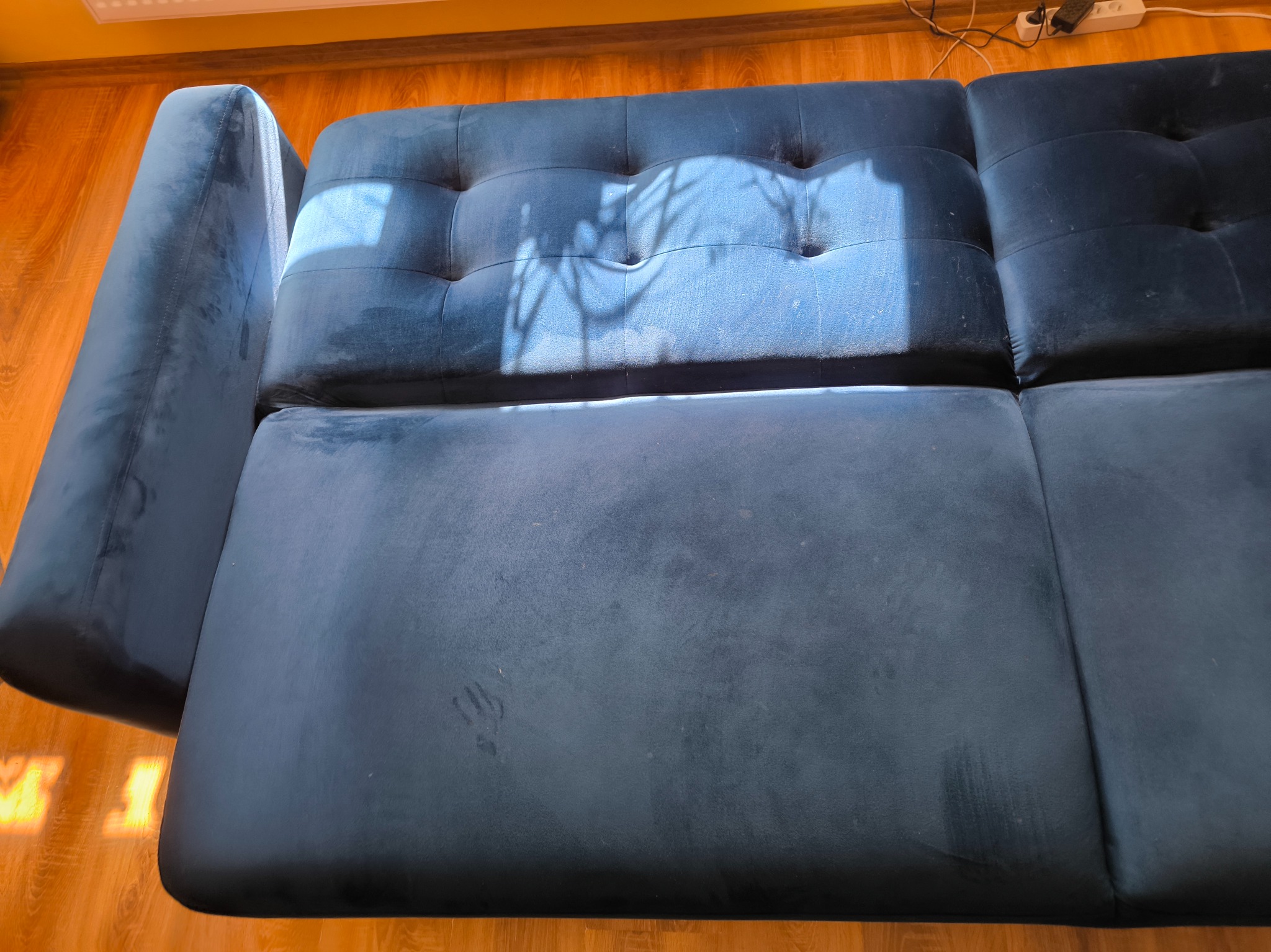 Ciemnoniebieska, welurowa kanapa z widocznymi plamami na siedzisku, oświetlona promieniami słońca. Widok z góry, ukazujący fakturę materiału i detale wykończenia.