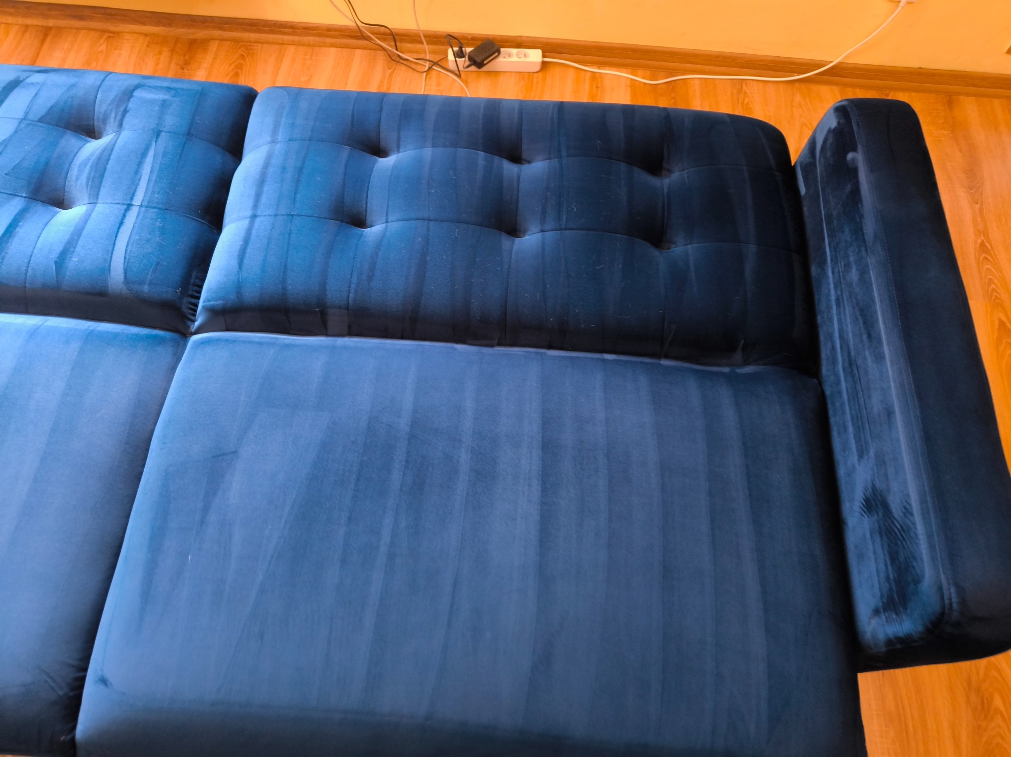 Granatowa, welurowa kanapa po czyszczeniu, widoczne ślady po praniu ekstrakcyjnym. Jasne smugi na ciemnej tapicerce. Widok z góry na drewnianą podłogę.