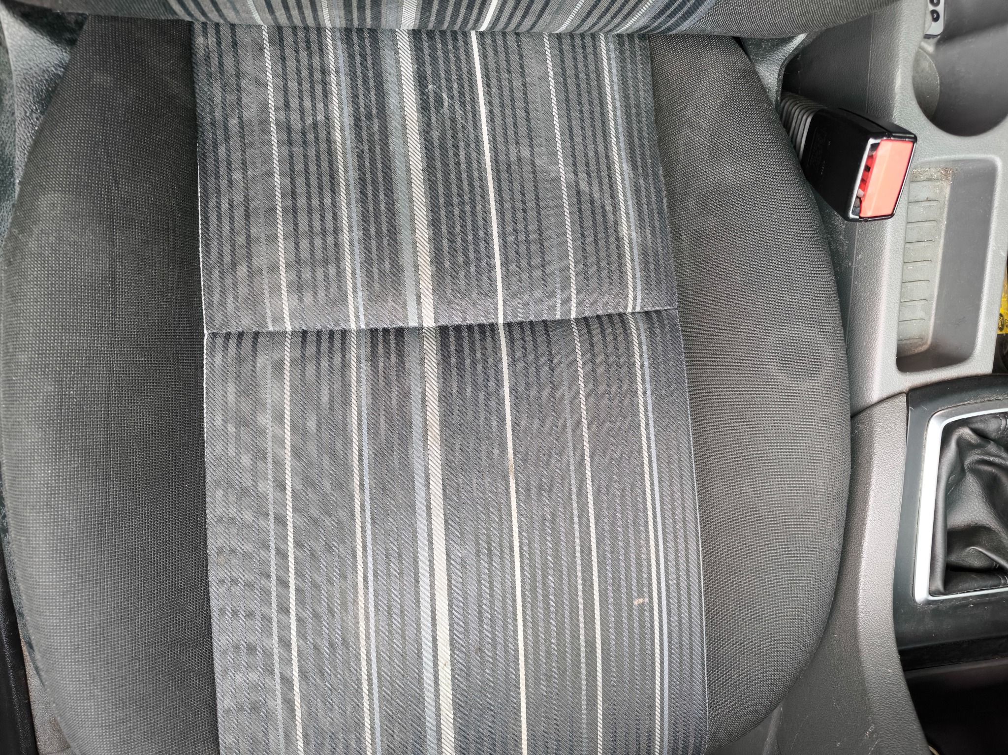 Szare, materiałowe siedzenie samochodowe z pasami w odcieniach szarości i bieli, widoczny fragment konsoli środkowej i zapięcie pasa bezpieczeństwa.