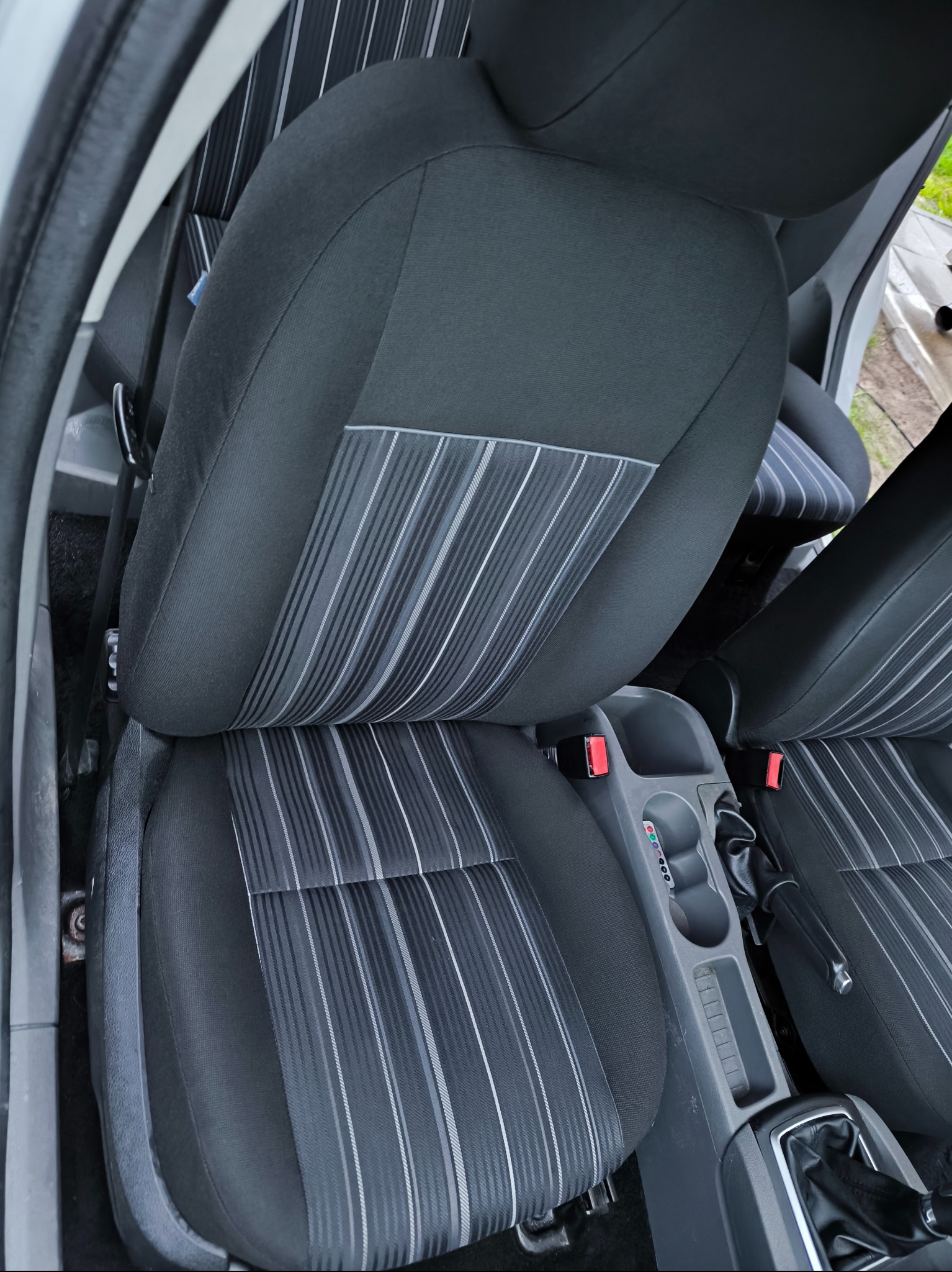 Czarne fotele samochodowe z jasnymi paskami, widoczne elementy konsoli środkowej i dźwignia zmiany biegów. Ujęcie z góry, wnętrze pojazdu.