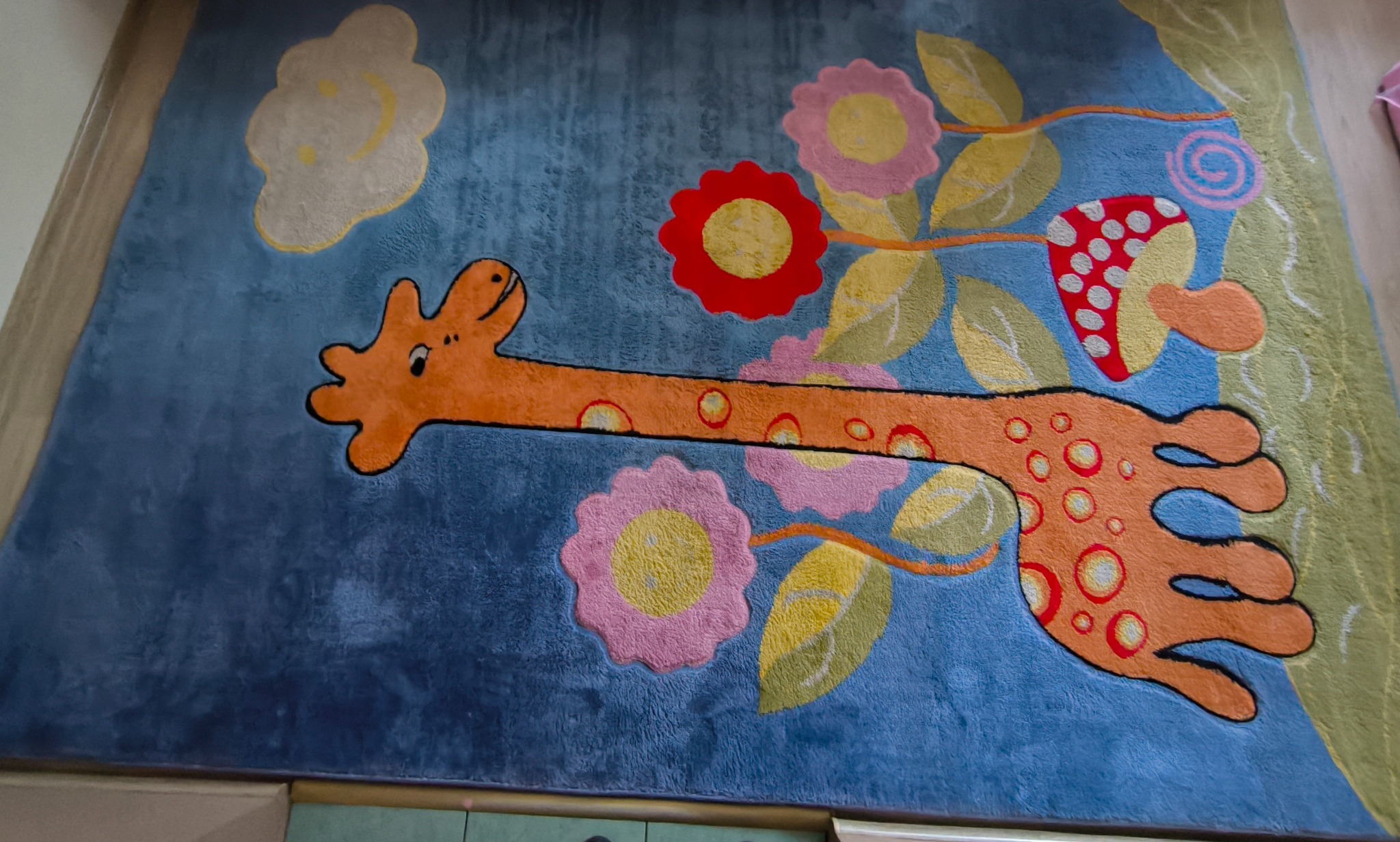 Niebieski dywan dziecięcy z motywem żyrafy, kwiatów i grzyba. Żyrafa ma pomarańczowe plamy z czerwonym obrysem. Dywan leży na podłodze, widoczny fragment ramy.