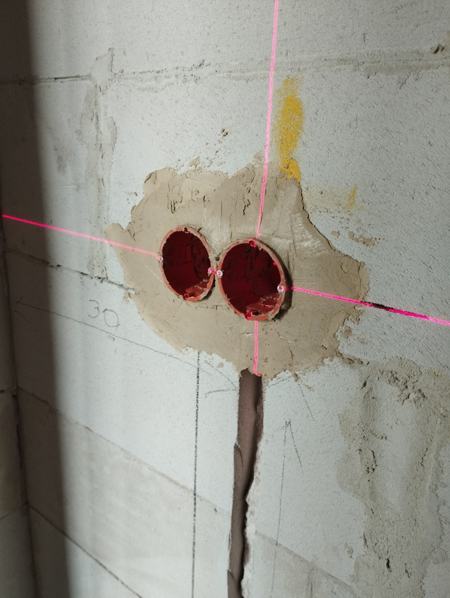 Dwa czerwone puszki elektryczne zamontowane w szarej ścianie, wyznaczane przez różowe linie lasera krzyżowego. Widoczne ślady po ołówku i zaprawa murarska.