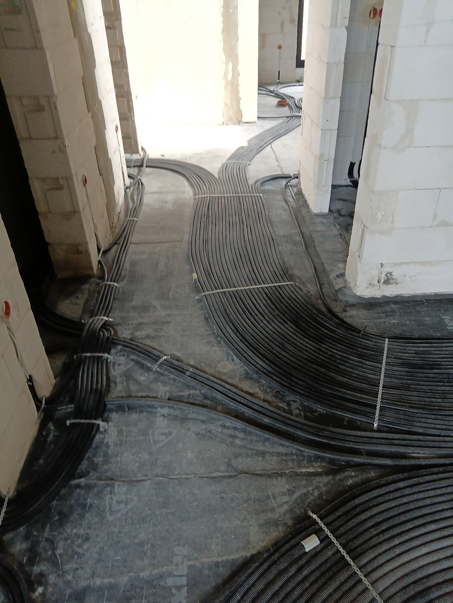 Kable elektryczne w peszlach ułożone na podłodze w surowym stanie budynku. Widoczne białe ściany z betonu komórkowego. Instalacja przed zalaniem wylewką.