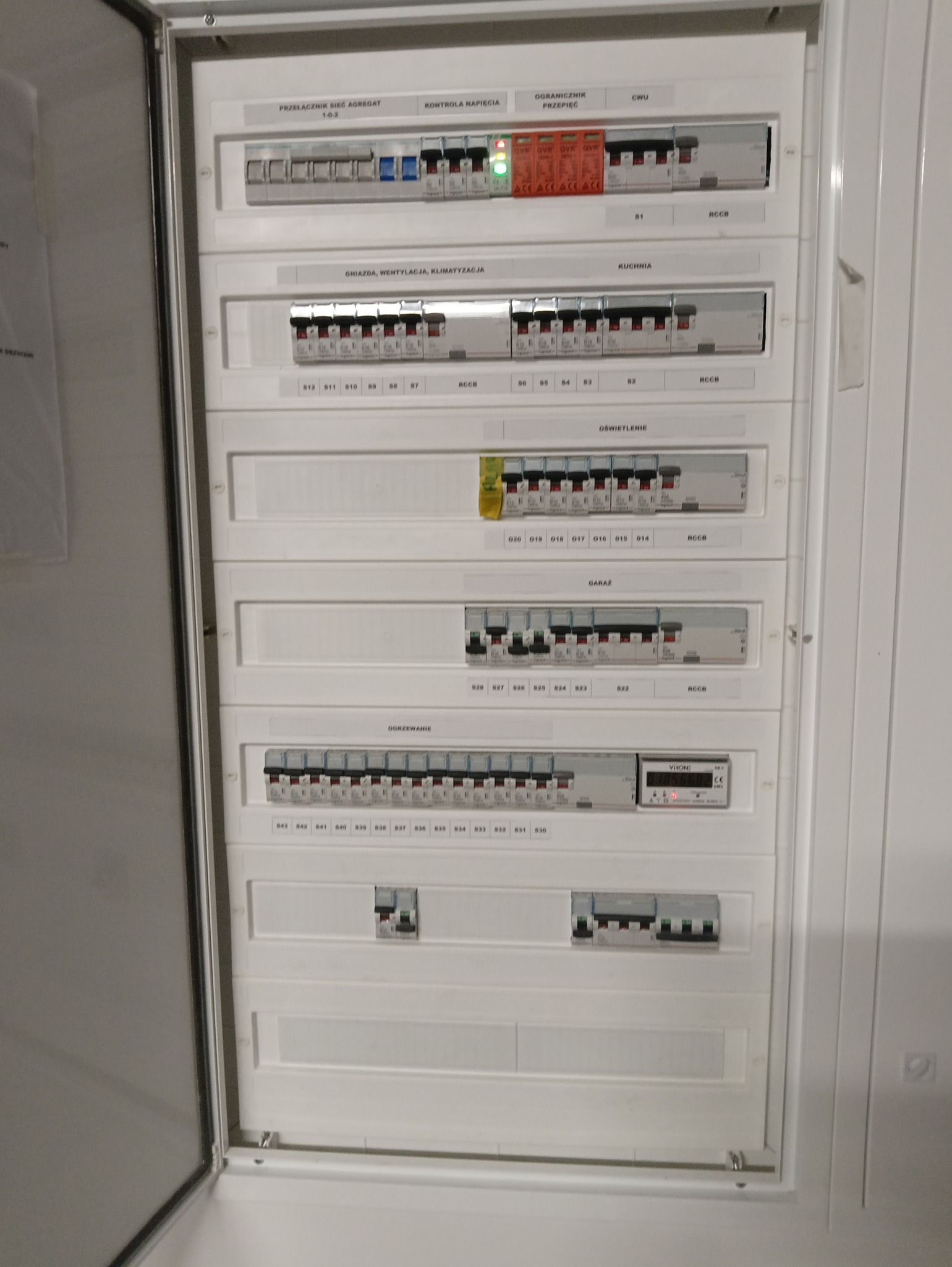 Rozdzielnica elektryczna z bezpiecznikami i opisami obwodów: oświetlenie, gniazda, wentylacja, klimatyzacja, kuchnia, garaż, ogrzewanie. Widoczne zabezpieczenia i kontrolki.