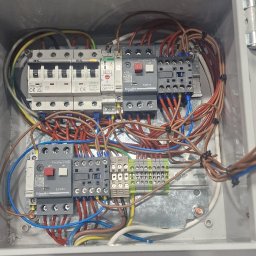 Łukasz Femiak - Otwarta skrzynka elektryczna z zamontowanymi bezpiecznikami, stycznikami i listwami zaciskowymi, widoczne różnokolorowe przewody oraz zapas końcówek kablowych w przezroczystym pudełku.