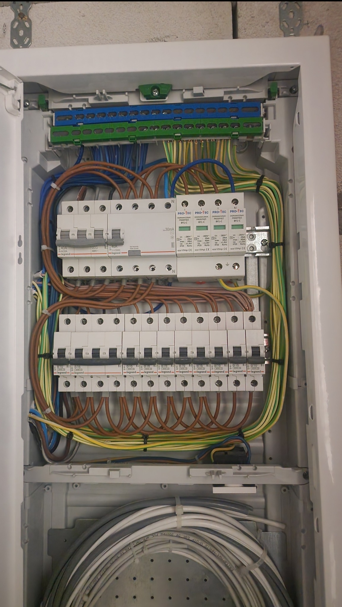 Otwarta skrzynka rozdzielcza z widocznymi bezpiecznikami Legrand, ogranicznikami przepięć Pro-Tec, przewodami o różnych kolorach i listwami zaciskowymi, całość zamontowana wewnątrz białej obudowy.