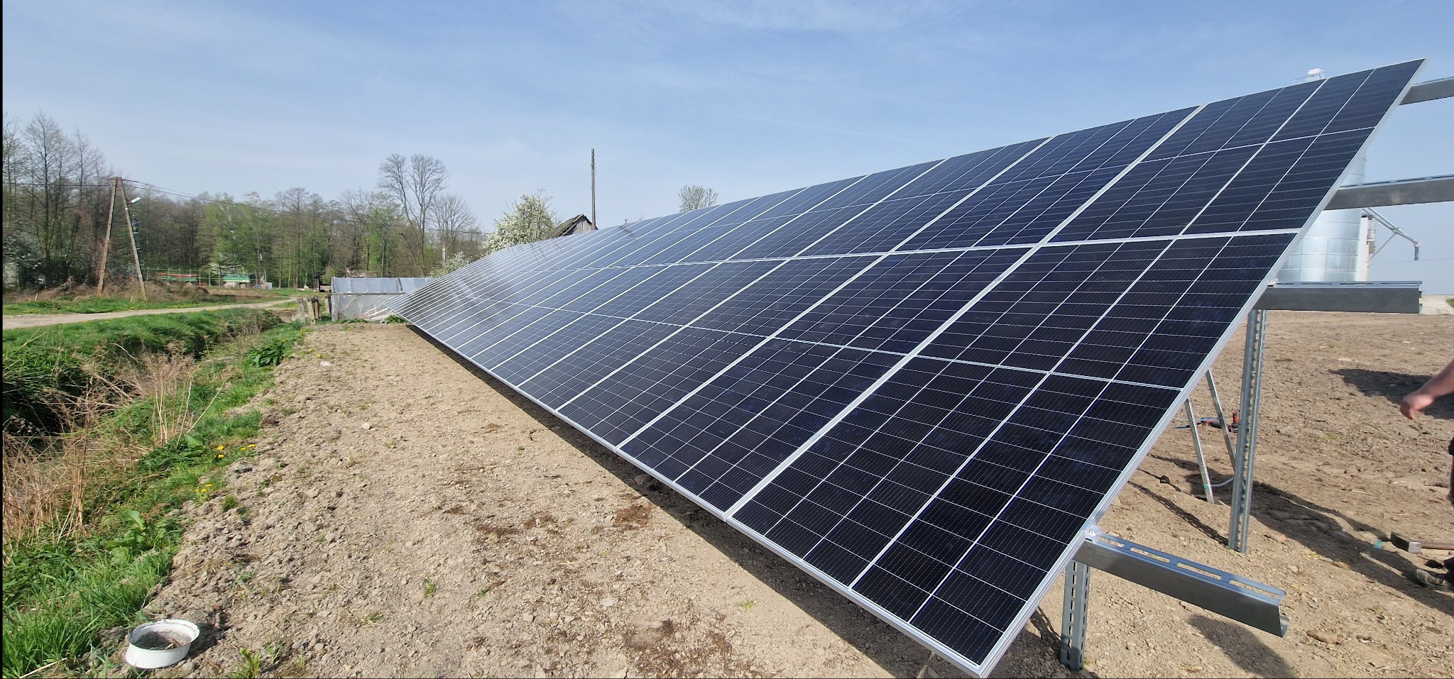Rząd paneli słonecznych na metalowej konstrukcji na tle wiosennego krajobrazu z drzewami i błękitnym niebem.