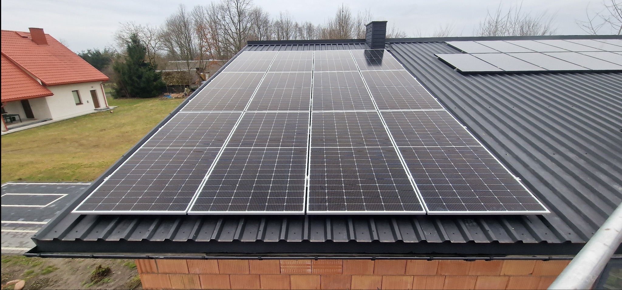 Panele fotowoltaiczne zamontowane na czarnym, blaszanym dachu domu jednorodzinnego z ceglaną elewacją, widoczne drzewa w tle.