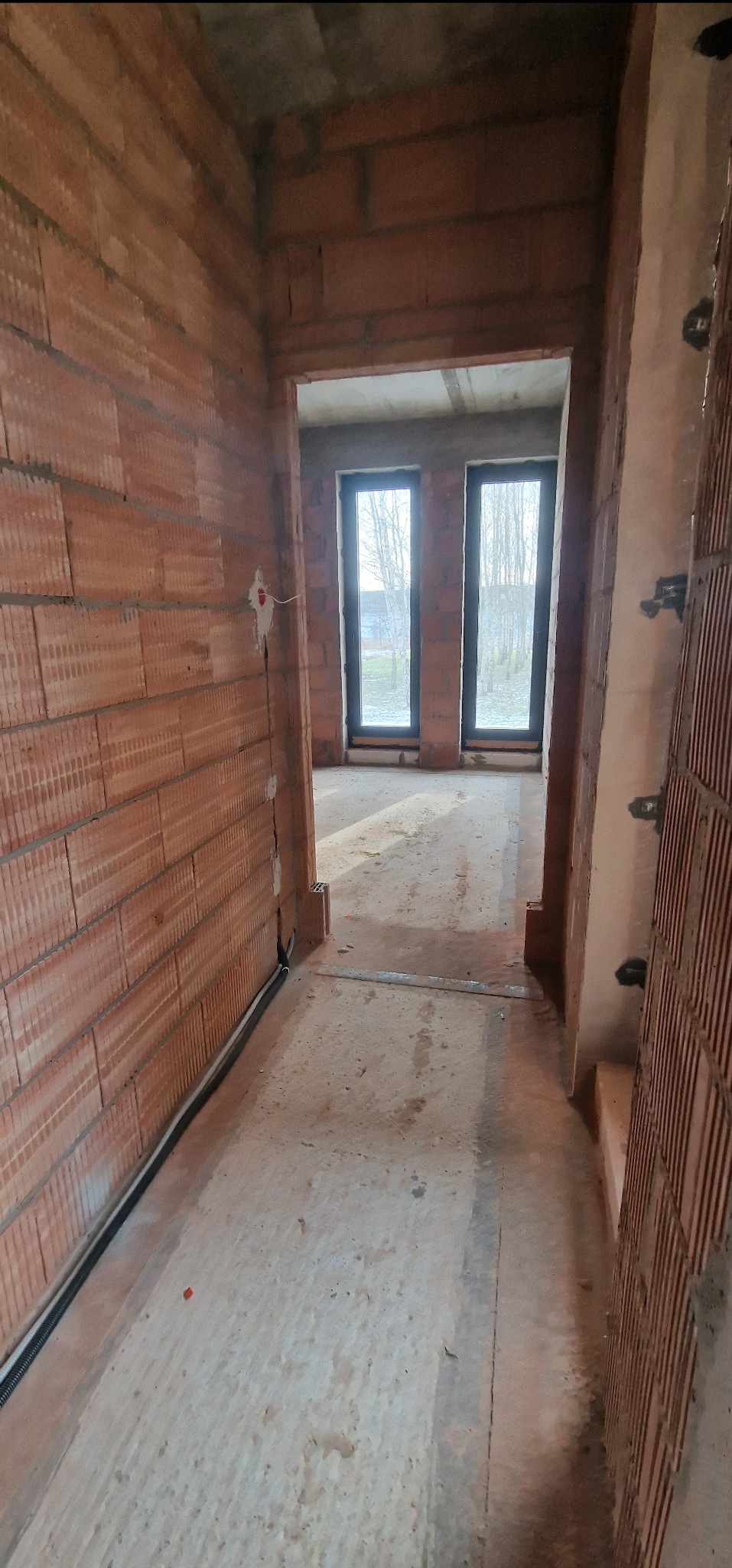 Wnętrze budynku w trakcie budowy: widoczne ściany z cegły, okablowanie elektryczne wzdłuż ściany i surowy betonowy sufit.