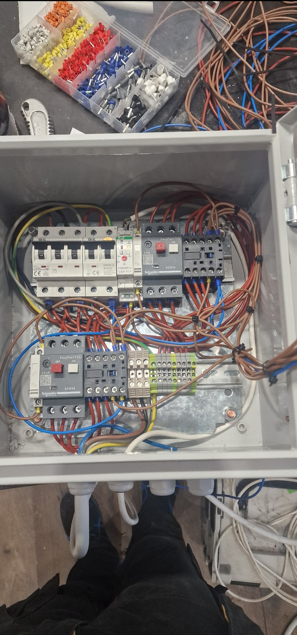 Otwarta skrzynka elektryczna z zamontowanymi bezpiecznikami, stycznikami i listwami zaciskowymi, widoczne różnokolorowe przewody oraz zapas końcówek kablowych w przezroczystym pudełku.