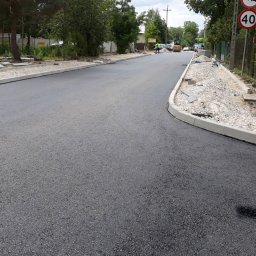 Geomas - Nowo położony asfalt na drodze z ułożonymi obrzeżami chodnikowymi, w tle maszyny budowlane i sterta kostki brukowej.