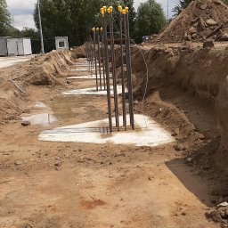 Geomas - Wykop z fundamentami punktowymi zalany betonem, wystające pręty zbrojeniowe zabezpieczone żółtymi zaślepkami, teren budowy z widocznym wykopem i stertą ziemi w tle.
