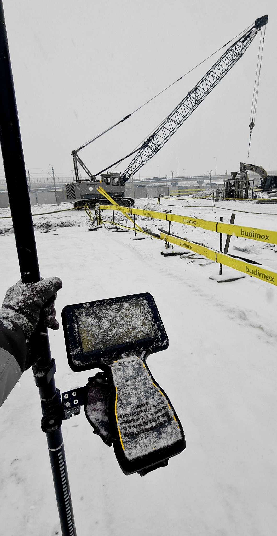 Praca geodezyjna w zimowych warunkach: kontroler GPS pokryty śniegiem, w tle żuraw budowlany i wykop zabezpieczony taśmami.