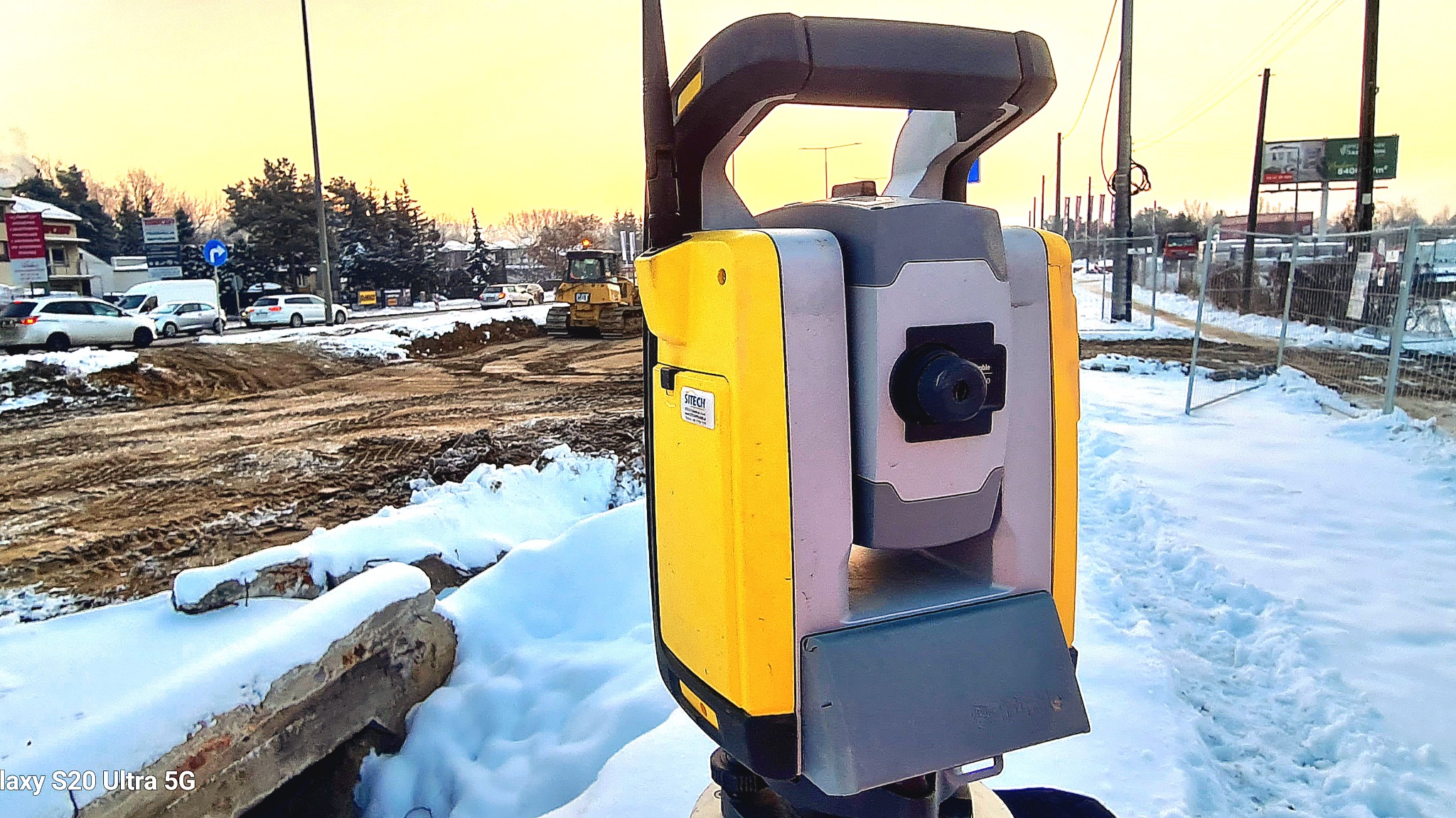 Żółto-szary tachimetr na statywie, w tle plac budowy z koparką i śniegiem, zimowa sceneria.