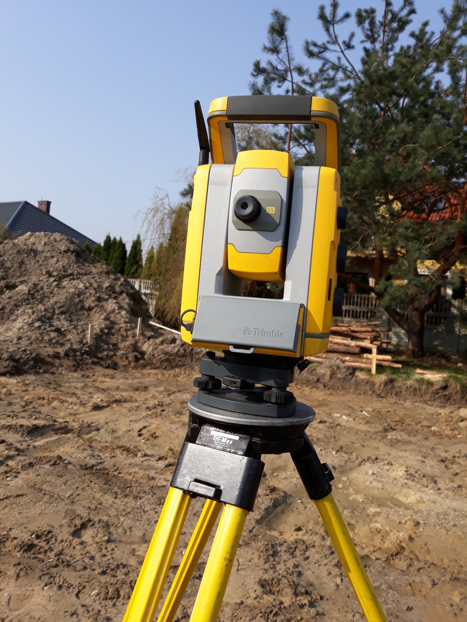 Żółto-szary tachimetr Trimble na statywie, używany do pomiarów geodezyjnych w Lublinie, z widocznym stosem ziemi w tle i fragmentem zabudowy.