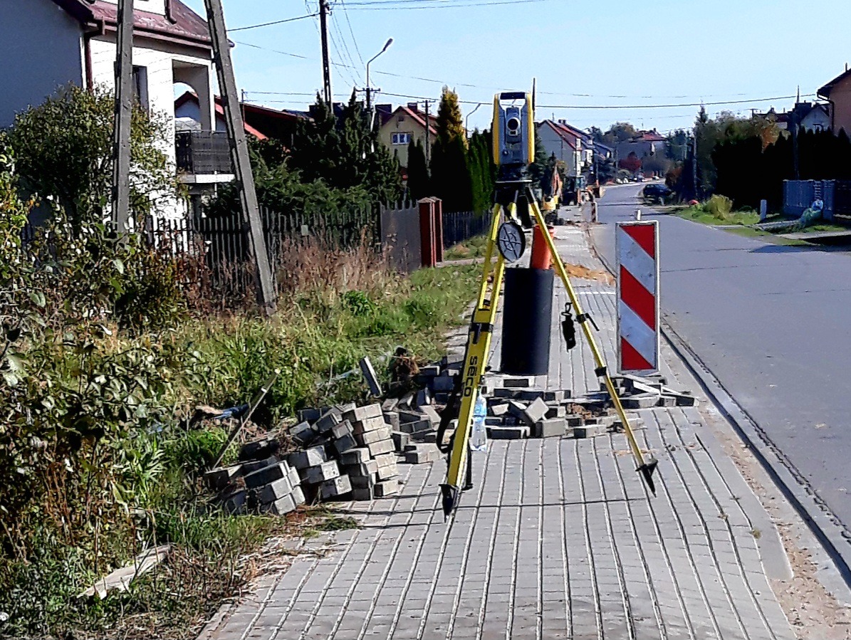 Żółty teodolit ustawiony na chodniku z kostki brukowej, obok sterta kostki i znak drogowy, w tle zabudowa jednorodzinna i prace ziemne.