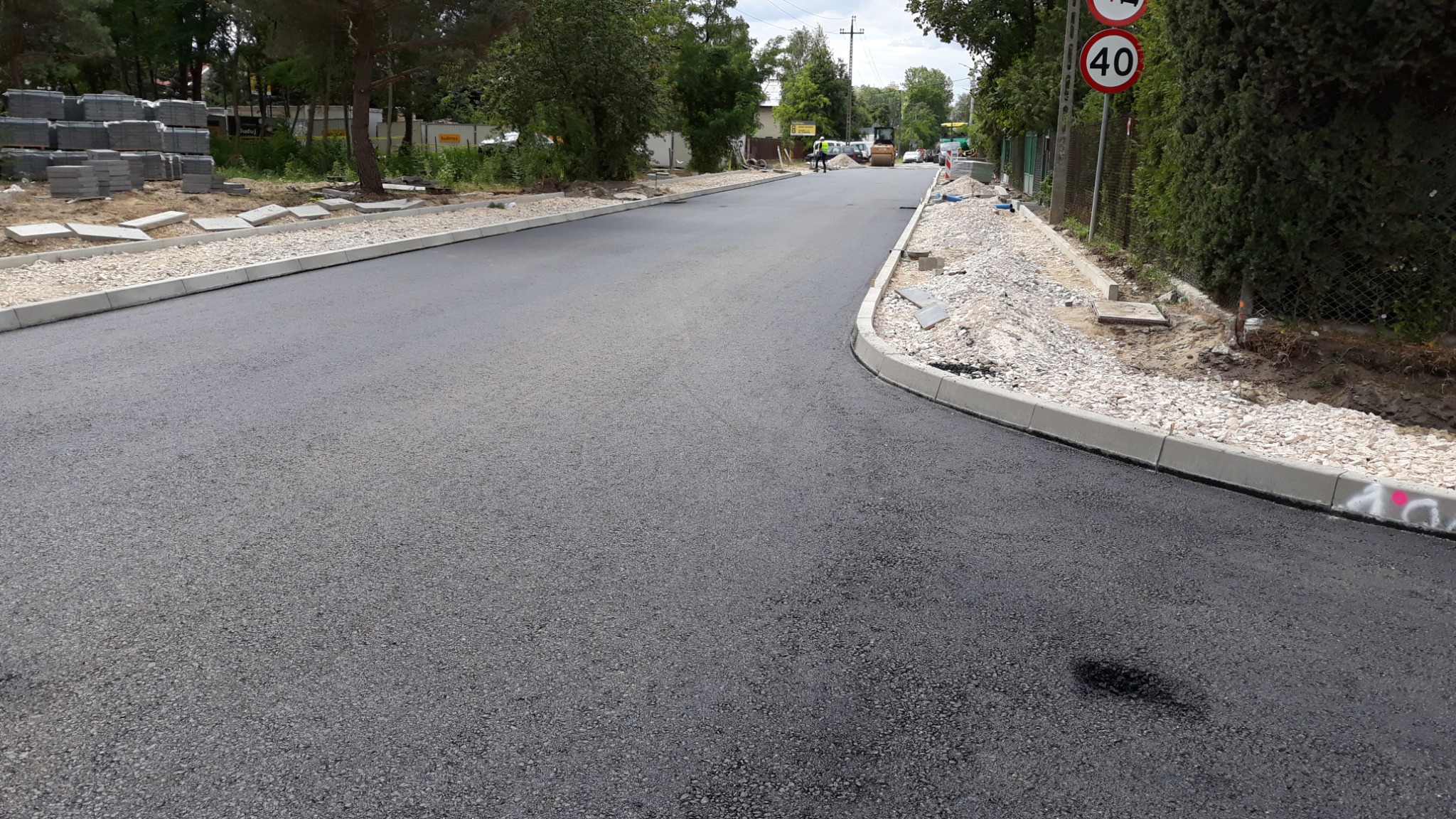 Nowo położony asfalt na drodze z ułożonymi obrzeżami chodnikowymi, w tle maszyny budowlane i sterta kostki brukowej.