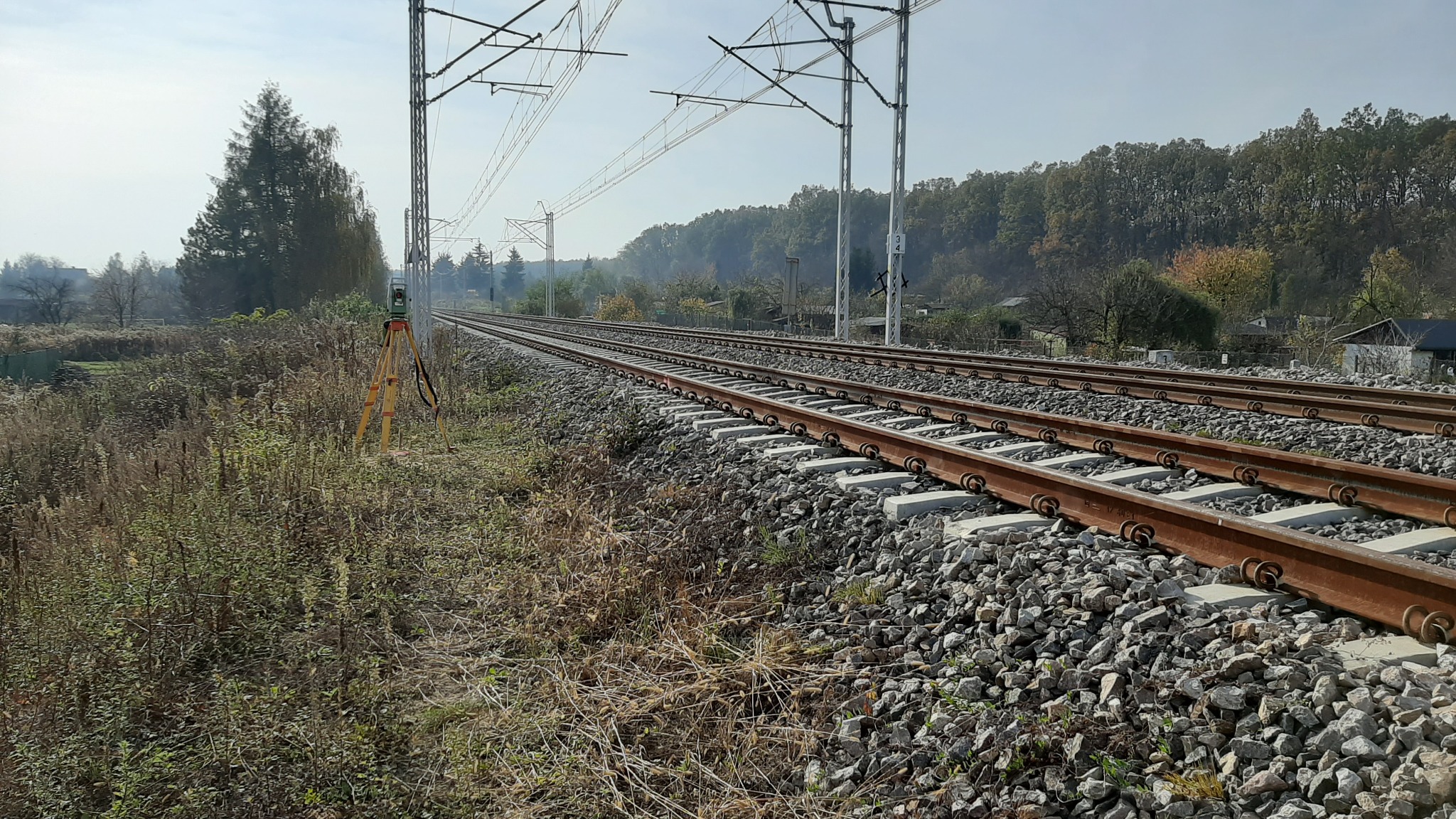 Pomiarowy instrument geodezyjny ustawiony na statywie obok torów kolejowych, w tle widoczne drzewa i słupy trakcji elektrycznej.
