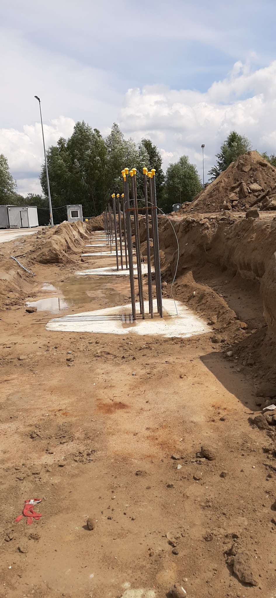 Wykop z fundamentami punktowymi zalany betonem, wystające pręty zbrojeniowe zabezpieczone żółtymi zaślepkami, teren budowy z widocznym wykopem i stertą ziemi w tle.