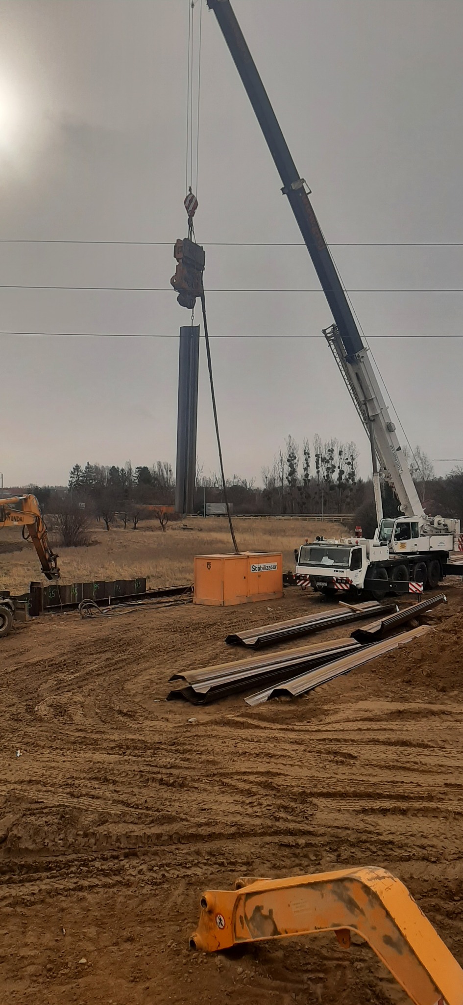 Żuraw przenosi stalowy profil w pionie na placu budowy, widoczny pomarańczowy generator, koparka Hyundai i ułożone profile stalowe na ziemi.