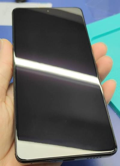 Dłoń trzyma nowoczesny smartfon z czarnym, błyszczącym ekranem, widoczne odbicia światła, tło niebiesko-turkusowe.