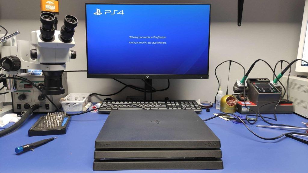 Stanowisko serwisowe z mikroskopem, lutownicą i konsolą PlayStation 4 Pro na niebieskim blacie, ekran monitora wyświetla komunikat powitalny.