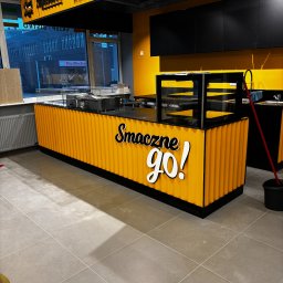 Aura Tek Sp. z o.o. - Wnętrze lokalu gastronomicznego w trakcie sprzątania: żółta lada z napisem 'Smaczne GO!', czarne szafki, mop i wiadro na szarej podłodze z płytek.