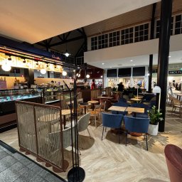 Aura Tek Sp. z o.o. - Wnętrze kawiarni z widocznym barem z ciastami i lodami, stolikami z niebieskimi krzesłami i roślinami w donicach, oświetlone jasnym światłem.