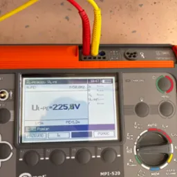 Miernik Sonel MPI-520 z podłączonymi przewodami pomiarowymi (czerwony i żółty) wyświetlający wynik pomiaru napięcia UL-PE = 225,8V na ekranie LCD.