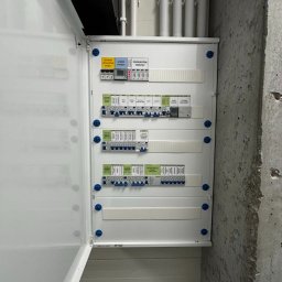 Aura Tek Sp. z o.o. - Otwarta, biała szafka rozdzielcza z bezpiecznikami i licznikiem energii, zamontowana na surowej, betonowej ścianie. Widoczne rury instalacyjne u góry.