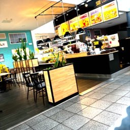 Aura Tek Sp. z o.o. - Kawiarnia z drewnianymi donicami i czarnymi lampami wiszącymi nad ladą. Turkusowa ściana w tle. Ustawienie stolików i krzeseł. Czarny blat.